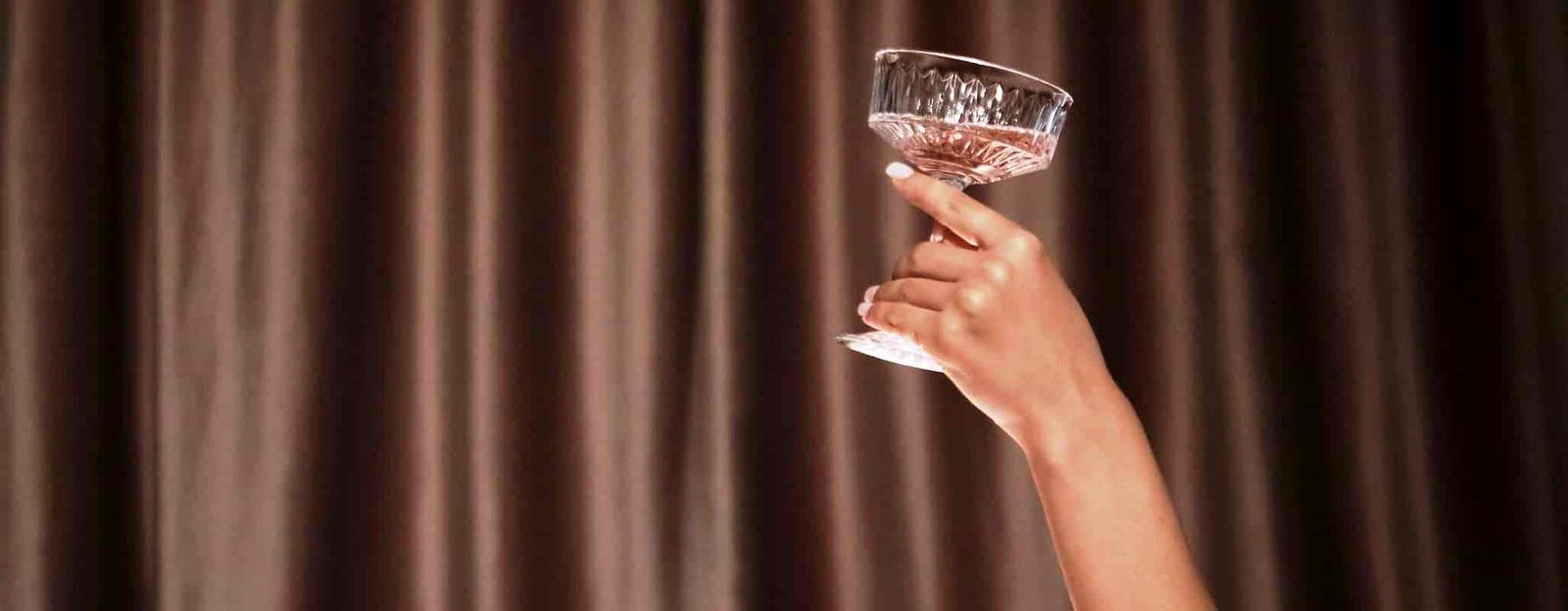A female escort arm extending from a luxury white bathtub, holding up a crystal coupe glass with pink champagne in a premium Delhi hotel suite.